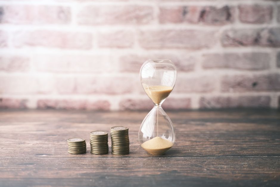A sequence of four neatly stacked piles of silver coins arranged in increasing order from left to right, positioned on a wooden surface indoors. To the right of the coins, an hourglass filled with bei