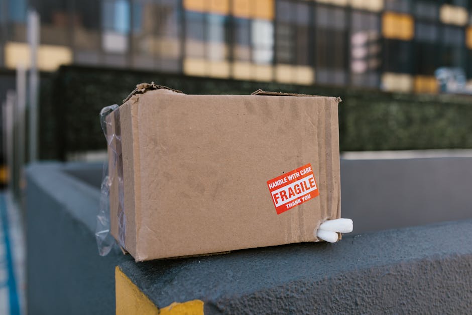A small rectangular cardboard box sealed with clear packing tape sits on the edge of a dark grey concrete surface outside a building during a home relocation process. The box features a red and white