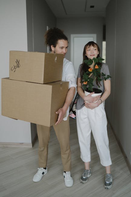 A man with dark hair and a beard, wearing a white t-shirt and blue jeans, is inside a room filled with numerous stacked cardboard boxes of various sizes, some sealed with packing tape and others open 